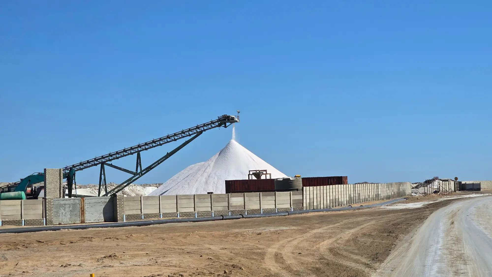 A white conical stockpile growing under a stacker conveyor in a walled open yard